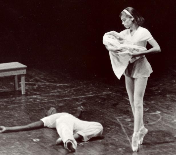 这张档案图片(黑白)描绘了一场芭蕾舞表演，舞者摆出了中间姿势, 脚尖站立，一条腿微微抬起, 抱着一个暗示婴儿的包裹. 她低头看着躺在她旁边地板上的一个舞者，创造了一个戏剧性的场景. 在后台, 在空荡荡的舞台上放着一张简单的木凳, 突出了舞者之间赤裸裸的情感时刻